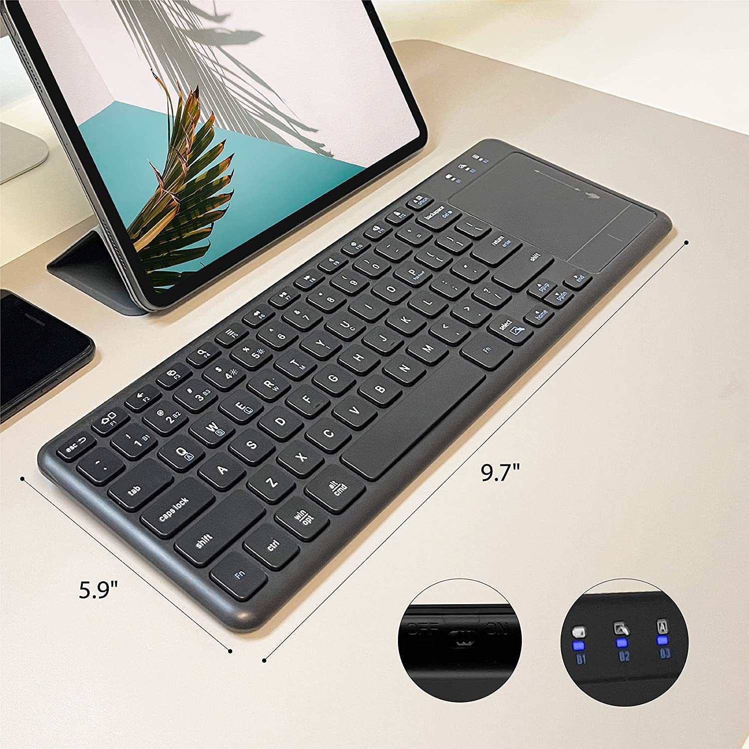 A slim black wireless keyboard with a trackpad is shown on a light beige desk next to a tablet and phone. White dimension lines indicate the keyboard's measurements: 9.