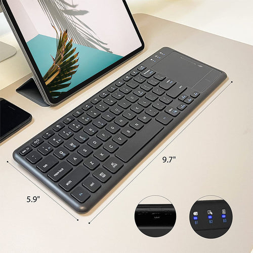 A slim black wireless keyboard with a trackpad is shown on a light beige desk next to a tablet and phone. White dimension lines indicate the keyboard's measurements: 9.